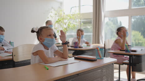 children-in-the-classroom-at-school-in-masks-sit-in-the-classroom-and-answer-the-teacher's-questions-by-raising-their-hands-in-slow-motion.-Lessons-during-the-pandemic-at-school